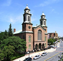 A brick church with two tall, symmetric steeples is seen in front of a city street, to the right of a wooded park.