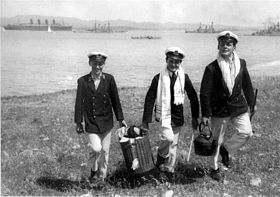 Black-and-white photo of three young men wearing World War I midshipmen uniforms carrying supplies for a picnic on a beach with three ships in the background.