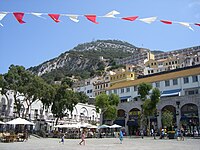 A broad, tree-lined sguare with shops and cafés on the perimeter and tiers of buildings on a slope leading up to the Rock of Gibraltar in the background