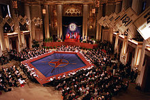 A long red table makes a pentagon around a blue floor with the NATO compass logo, while many rows of people in suits sit on all sides.