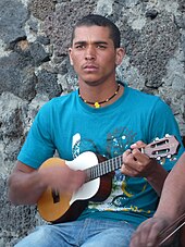 A young man holds a small four-stringed instrument and strums.