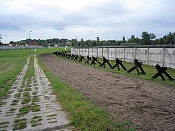 Strip of bare ploughed earth flanked by a concrete road on one side and a row of barricades and a fence on the other side, with buildings visible in the far background.