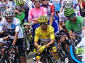 Three cyclists waiting with their bicycles in front of several others or more, with each of the three wearing white, yellow and green jerseys
