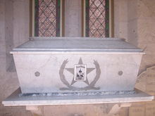 A white marble coffin sits on a ledge in front of stained glass windows. On the front of the coffin is a large 5-pointed star. Engraved within the star are the words "Texas Heroes" and small images of three men.
