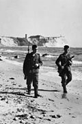 Two armed men, one with a rifle with a bayonet attached and the other carrying a submachine gun, walking along the water's edge below a line of cliffs.