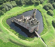 An aerial view of a stone building with a triangular plan. It is surrounded by a ditch filled with water.