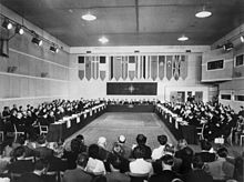 A long rectangular room with multiple rows of seated individuals on each side, and flags hanging at the far end.