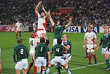 Two rows of opposing players, green to the fore, white behind, each aiding a jumping player from their team by lifting him towards an off-picture ball travelling overhead