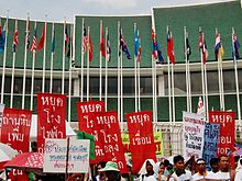 Many people holding signs in front of a building with green layered roofs; many national flags on flag poles lined in two rows in front of the building