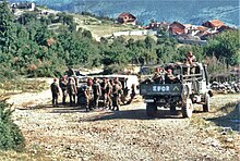 Three trucks of soldiers idle on a country road in front of trees and red-roofed houses. The rear truck has KFOR painted on is back.