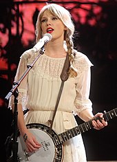 Taylor Swift performing live on a banjo, wearing a beige blouse and pigtails