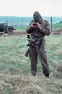 Armed East German guard standing in a grassy field taking a photograph of the photographer. A border fence and a truck are visible in the background, some distance behind the soldier.