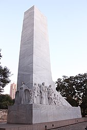 The rectangular base of a cenotaph. An angel is carved on one end. On the side are carvings of several men, shown wearing bucksin or 19th-century suits. Many hold guns or knives; at the far end, one operates a cannon.