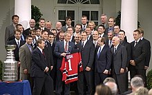 President George W. Bush receives the New Jersey Devils players in the White House doors. Bush holds the Devils jersey with the name Bush and the number 1. To the left of the crowd, the Stanley Cup sits on a table.