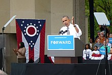 A black man in a white shirt speaking on stage behind a podium with the word "forward" and in front of a red, white, and blue Ohio state flag.