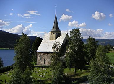 A little stone church with a little steeple on a wooden belfry sits in a green graveyard overlooking a lake and mountains.