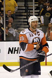 A Caucasian male wearing an orange, blue, and white jersey. Guerin is standing in a hockey rink, holding a hockey stick and has his right arm held against his stomach.