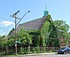 An ivy-covered building with a pointed roof and a small green dome with a cross seen from across a street, with a car parked in front. There is a chainlink fence around it and the ground slopes downwards towards its rear. A street sign at the corner reads "Colonie" and "North Pearl".