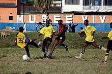 five men playing football one red and black striped jersey and four in yellow jersey