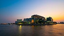 A large complex of buildings, most over ten storeys high, on the bank of a river; one bears a sign with the words "SIRIRAJ HOSPITAL"; another says "FACULTY OF NURSING"