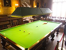 A full-size snooker table in a brightly-lit room with bookcases and a boardroom table in the background, all cordoned off at the right-hand side as part of an English country house display