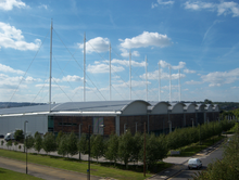 A Modern sporting facility. The building is roughly rectangular in shape and is quite modern, with a wooden clad design. It is surrounded by greenery and has several tall, white spikes on its roof.