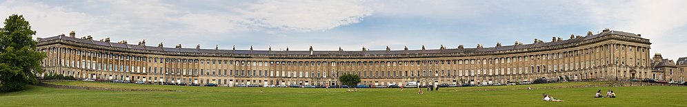 Wide image of a symmetrical semicircular terrace of yellow stone buildings. Grass in the foreground.
