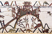 A section of an embroidered cloth showing a castle on a hilltop being defended by soldiers with spears while two soldiers in armour are attempting to set fire to the palisade