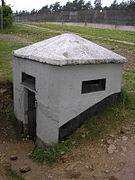 A square white half-buried concrete bunker with a concrete broach roof and rectangular horizontal loopholes. A tall fence is visible in the background.