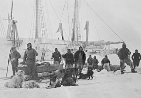 A group of men pose on the ice with dogs and sledges, with a ship's outline visible in the background