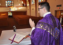 A man wearing purple vestments and standing at an altar uses a mobile phone camera to record himself. Empty pews are visible in the background.