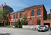 A wide two-story brick building with a hipped roof and gently arched windows, some of which have been bricked in. Across the top is a sign saying "Albany Pump Station." There is a taller building on the left.