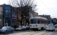 A white single-car trolley in street running. On street parking combined with double track on a two-lane street leaves limited room for automobile maneuverability.