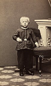 an unsmiling fair-haired child stands upright, his left hand resting on a stool, in front of an ornate fireplace.