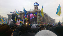 Dozens of blue and yellow Ukrainian flags are held aloft in a wide crowd watching large screens on a square building.