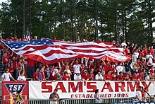 U.S. soccer fans, dressed in red, cheer in bleachers as they hold a large U.S. flag over themselves at a soccer match.