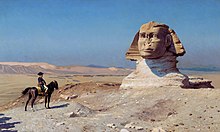 Person on a horse looks towards a giant statue of a head in the desert, with a blue sky