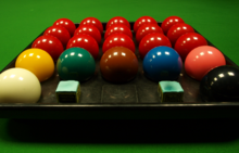 Close-up view of an open snooker ball box with three rows of five red balls to the rear, one row of colour balls towards the front, a white ball to front left corner, a black ball to front right corner, and two chalk cubes at the front between the white and black balls