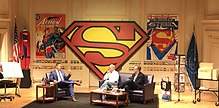 Three men seated onstage, flanked by Superman material