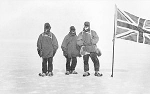 Tres hombres con ropa pesada hacen fila sobre una superficie helada, junto a un asta de bandera desde la que ondea la bandera del Reino Unido de Gran Bretaña e Irlanda