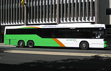 Light green, orange, and white bus stopping in front of multi-story building.