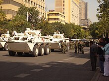 A caravan of white armoured personnel carriers rolling through a city street, with soldiers carrying body shields marching alongside. Several civilians are standing by on the pavement.