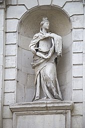 A photograph of a white statue of a woman clothed in a long robe looking to the right and placing her left hand on her chest in a white archway in a wall