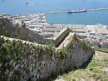 View of a stone wall descending a steep slope, with a harbour in the background