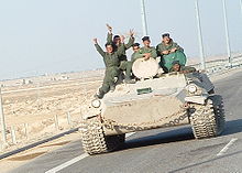Six uniformed soldiers waving from an armoured vehicle on a highway