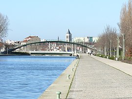 Canal Saint-Denis พร้อมสะพานชิงช้า สะพานลอยสำหรับคนเดินที่นำไปสู่ ​​Stade de France และ Basilique Saint-Denis ในพื้นหลัง