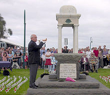 Photo of an ANZAC memorial with an elderly man playing the bugle. Rows of people are seated behind the memorial. Many small white crosses with red poppies have been stuck into the lawn in rows on either side of the memorial.