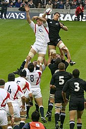 The All Blacks and England contesting a line-out. Both sets of forwards lined up wearing white and black respectively, with a player from each side at the rear of the line out being lifted by their teammates while both reaching for the ball.
