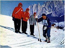 A man in blue clothes with a number "33" cross-country skiing in front of three other men.