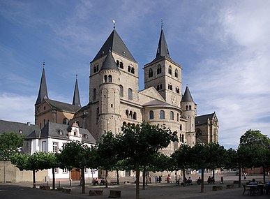 A huge cathedral with numerous towers, both square and round, rises above a town square where people are sitting in the shade of clipped trees.